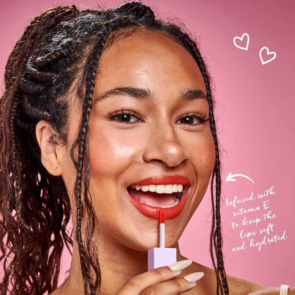 A close up of a model applying one of the matte liquid lipsticks to her lips in front of a pink background. An arrow with text saying it includes Vitamin E to keep the lips soft and hydrated.