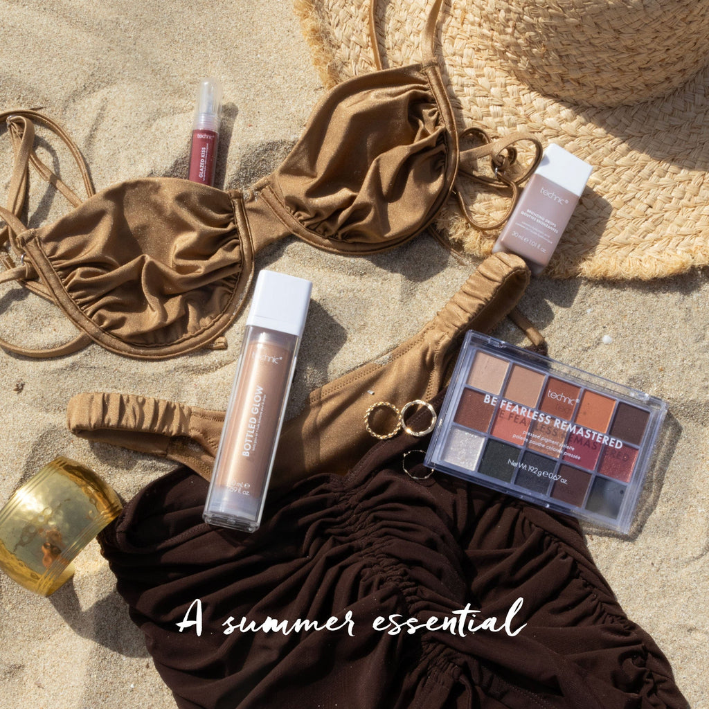 A bikini set and a sun hat on the sand with bottled glow, bronzing drops, a coloured lip gloss and an eyeshadow palette around it.