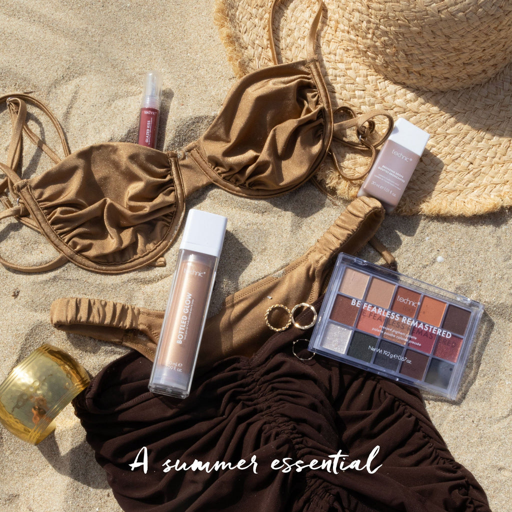 A bikini set and a sun hat on the sand with bottled glow, bronzing drops, a coloured lip gloss and an eyeshadow palette around it.